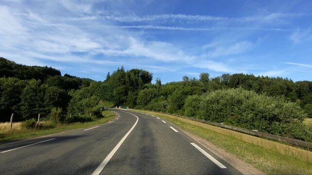 Curving rural road winding through lush green forest under a bright blue sky in the Morvan region of France. Peaceful summer landscape ideal for travel and nature concepts.