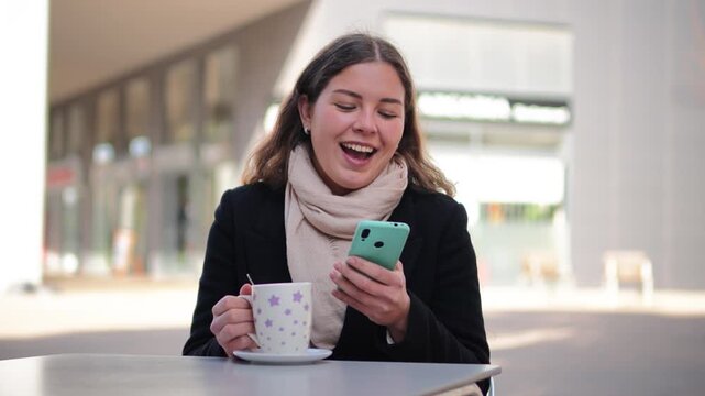 A young woman is sitting at an outdoor cafe, smiling as she texts on her smartphone while sipping a warm beverage. The bright atmosphere enhances her joyful experience in this delightful moment.