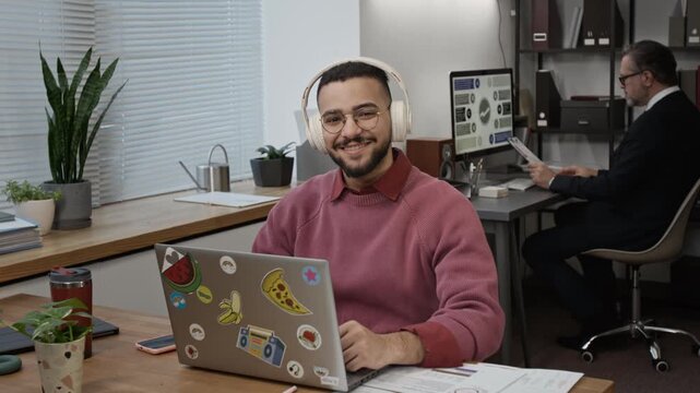Portrait of young male data analyst wearing pink sweater and beige headphones smiling for camera working on grey laptop decorated with stickers in office, with formally dressed man in background