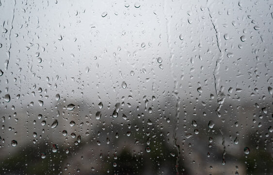Rain drops on window glass during cloudy rainy weather with blurred city view in the background. Concept of autumn weather, sadness, melancholy, storm, climate, and urban rainy day background.