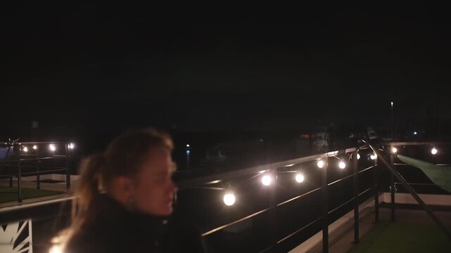 A person in soft focus sips a drink on a dark night, surrounded by warm, glowing string lights on a balcony overlooking a distant marina. Evokes a relaxed, cozy evening atmosphere
