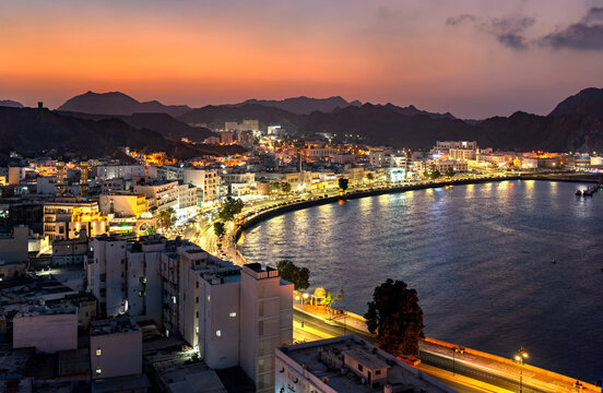 A stunning panoramic view of the illuminated Mutrah Corniche and harbor area at sunset, showcasing the coastal architecture and mountainous backdrop in Muscat, Oman.