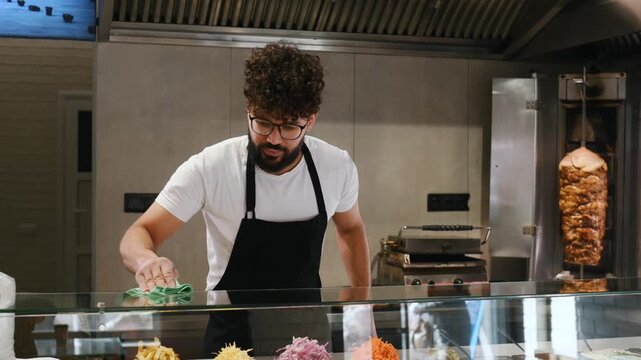 Male vendor with curly hair and glasses wipes down a glass display case in a restaurant, showcasing grilled meat and various food items in the background