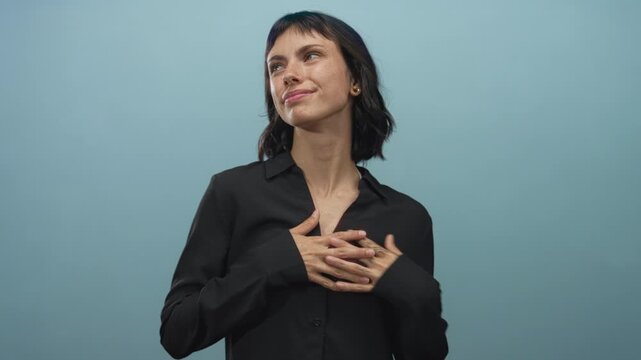 Young hispanic woman clutching chest in light blue studio, looking upward with gentle smile; serenity.