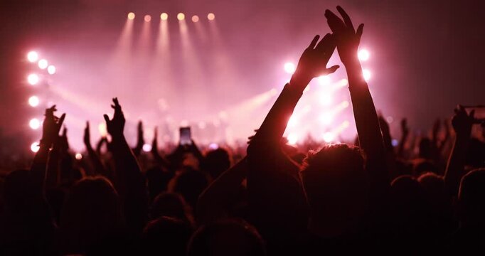 A large group of people raises their hands in applause during a performance at a concert hall. Warm pink and red stage lights illuminate the cheering crowd.