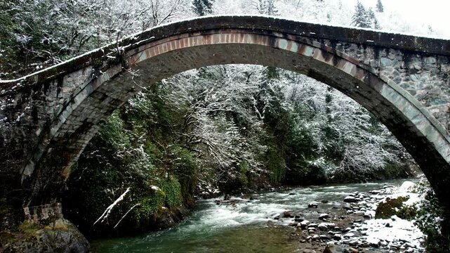 Gentle snowfall over a historic stone arch bridge crossing a clear mountain stream in Ayder Plateau, Rize, Turkey. Snow-covered forest and flowing water create a peaceful winter nature landscape.