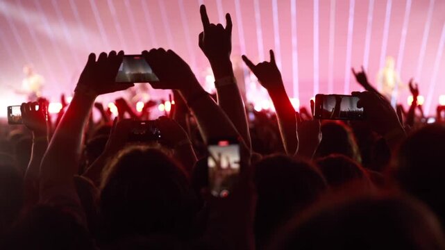 A large crowd waves their hands in unison during a live musical performance. Intense red stage lighting creates a dramatic and atmospheric visual effect.