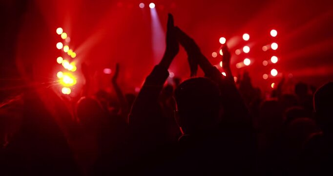 A crowd of fans raises their hands and claps in rhythm at a live music performance. Bright red and yellow spotlights create a vibrant atmosphere in the dark venue.