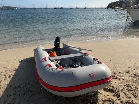 Mallorca, spain, february 20, 2026, gray inflatable dinghy with outboard motor beached on sandy shore as calm blue sea laps gently, sailboats anchored nearby