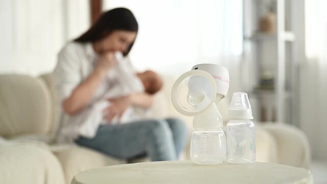 Breast pump and two baby bottles are positioned on a table, while a woman is breastfeeding her baby on a couch in a cozy living room