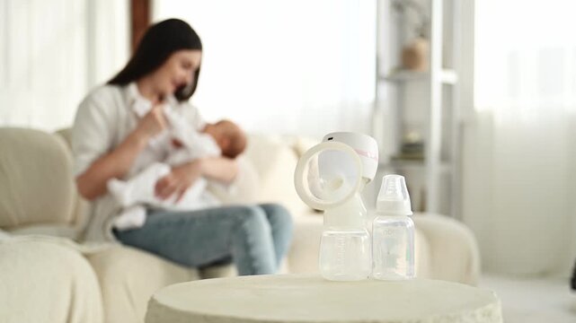 Breast pump and two baby bottles are positioned on a table, while a woman is breastfeeding her baby on a couch in a cozy living room