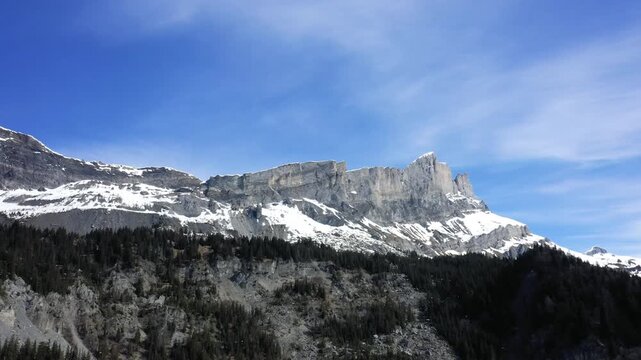 Dramatic snow dusted rocky cliffs of Tete a l ane rise above a dense pine forest under a clear blue sky. Majestic alpine landscape in spring.