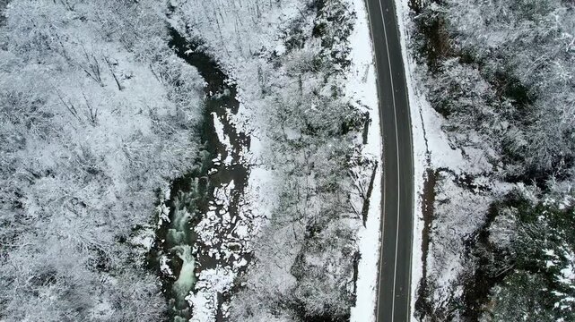 Top-down aerial view of a snowy forest landscape with a winding mountain road and flowing river in Rize Ayder, Turkey. Winter scenery with snow-covered trees creates a calm and peaceful nature atmosph
