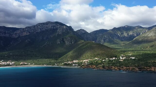 Scenic view of a coastal village with turquoise waters and lush green mountains near Monemvasia, Greece, under a dramatic cloudy sky.