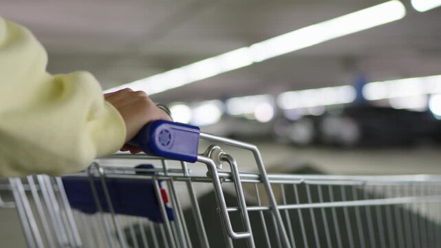 Close-up of a woman hand on the handle of a shopping cart, pushing it through a brightly lit underground parking garage. Shallow depth of field blurs the background