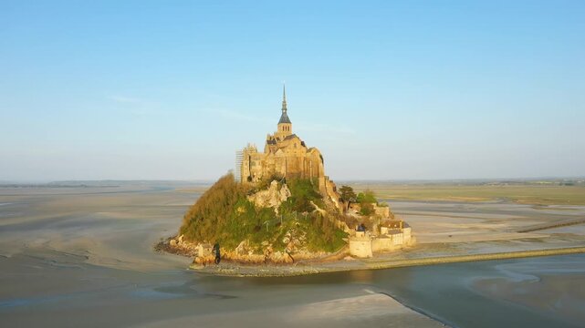 Scenic drone shot of Le Mont Saint Michel island and abbey surrounded by tidal flats in Normandy, France, under clear blue sky.