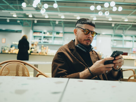 A man in a cafe holds a phone and watches the news wearing orange glasses