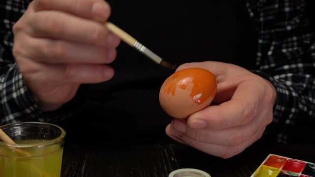 Person painting an orange design on an egg with a fine brush, surrounded by paint supplies and a glass of yellow dye on a dark wooden table