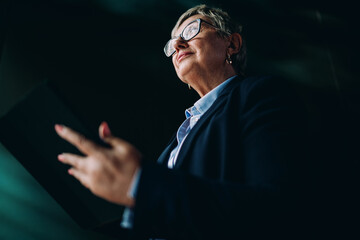 Low-angle view of senior woman holding tablet, looking determined. Strong visual message of modern leadership, authority, and digital confidence in a mobile world.