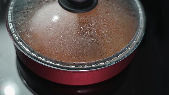 A close-up view of a pot lid with steam condensing on the glass. The pot sits on a stove, indicating a cooking process.