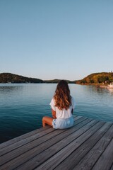 Fototapeta premium Female caucasian young sitting on a wooden dock facing a calm lake at sunset overlooking distant tree lined shore