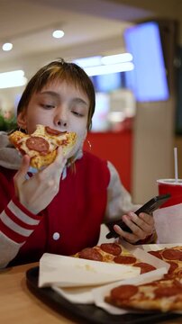 A teenage girl balances her love for pizza and social media, scrolling through her phone while savoring every bite, her attention fully captured by the online world.