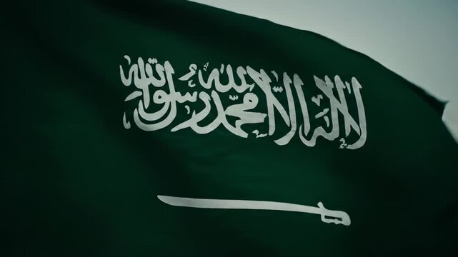 Close-up view of the national flag of Saudi Arabia, featuring the green field, white Shahada inscription, and a white saber, waving gently.