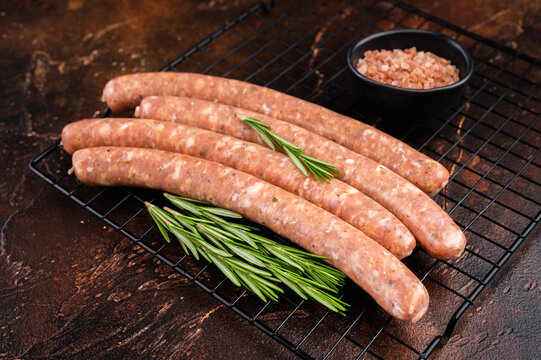 Raw fresh Chipolata sausages on metal grill rack with rosemary and coarse salt on dark rustic background. Uncooked pork meat links ready for grilling barbecue.