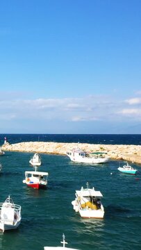 Small fishing boats float near a stone breakwater under a clear blue sky in the harbor of Koroni, Greece. Calm sea and bright daylight create a tranquil coastal scene.