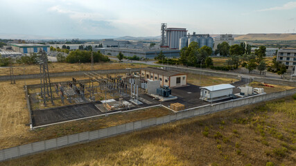 Obraz premium Aerial view features an electrical substation with transformers and metal structures in a fenced area, set against an industrial backdrop with large factory buildings and silos under a blue sky.