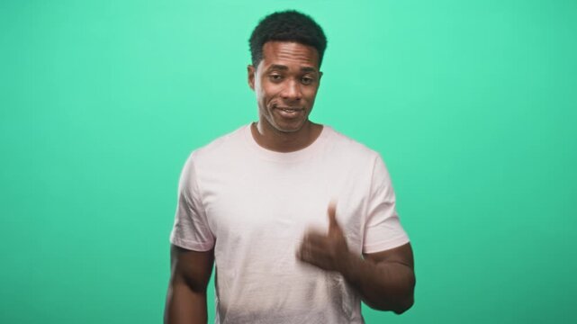 Man pointing thumb to side in studio with green backdrop, wearing pale pink tshirt and smirking with a shrug; playful.