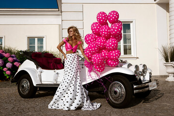 Glamorous Woman in Black and White Polka Dot Dress Posing with Vintage White Convertible Car, Pink Balloons and Roses outside a Luxury Palace Mansion