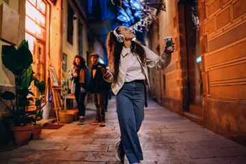 Smiling multiracial girl walking in narrow European street at night with coffee and phone, wearing...