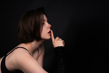 Obraz premium Woman in lingerie lying on stomach on black background making hush gesture with finger on lips, erotic concept, side profile, low key studio lighting.