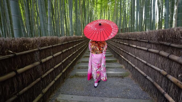 Woman wearing traditional kimono with red umbrella walking through the famous bamboo forest path in Arashiyama, Kyoto, Japan. Iconic travel destination showcasing Japanese culture and natural beauty.