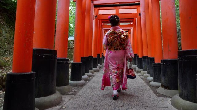 Asian woman wearing traditional kimono walking through the famous red torii gates at Fushimi Inari Shrine in Kyoto, Japan. Cultural travel destination showcasing historic Japanese heritage and Shinto 