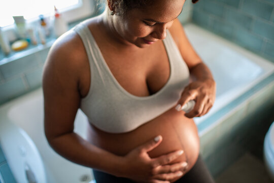 Pregnant woman applying belly lotion in home bathroom