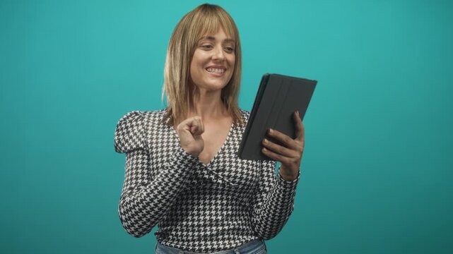 Woman taps tablet screen with index finger, smiling and holding device in turquoise studio setting; delight curiosity connection.