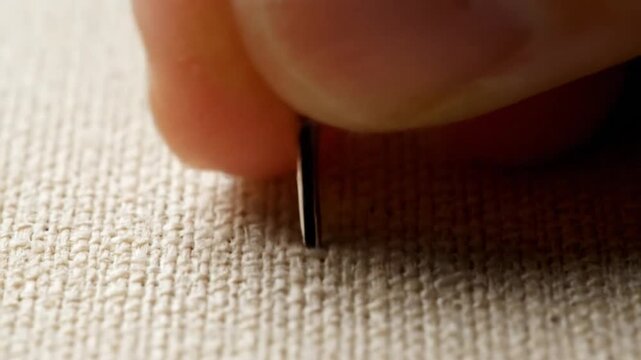 A hand uses a sewing needle to stitch through beige fabric, creating a loop; close-up view emphasizes the detailed threading process.