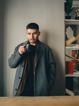 A man in a gray coat holding a black coffee mug against a wall in a cafe interior in spring. He looks at the camera with a short haircut.