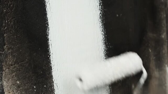 A person paints a textured wall with white paint using a roller brush, gradually covering a dark surface in a series of sequential movements, focusing on the painting process.