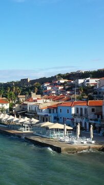 Colorful waterfront buildings and outdoor cafes with umbrellas line the promenade in Koroni, Greece, with a historic hilltop structure in the background under a clear blue sky.