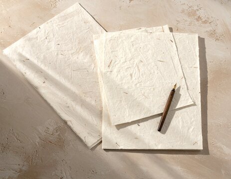 Hojas de papel artesanal y pluma sobre superficie texturada de adobe claro