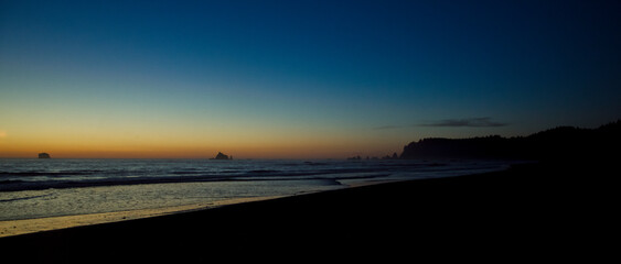 Ocean sunset over a dark sandy beach with silhouetted sea stacks and coastline, twilight sky shifting from deep blue to vibrant orange for a calm, peaceful seascape © NJF