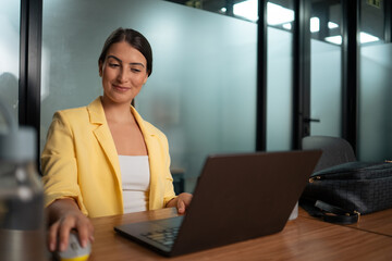 Professional woman in yellow blazer working on laptop at wooden desk in modern corporate office with glass walls