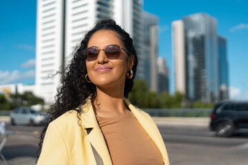 Young professional woman with sunglasses and yellow blazer standing on urban street with modern skyscrapers in background