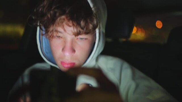 Close-up of a teen boy in a hoodie in a dark car at night. His face is lit by his phone screen as he looks down, focused or sad. Out-of-focus city lights are visible in the background