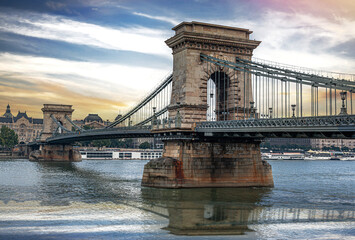 Obraz premium The Szechenyi Chain Bridge spans the Danube River in Budapest under a dramatic sunset sky with historic architecture in the background.