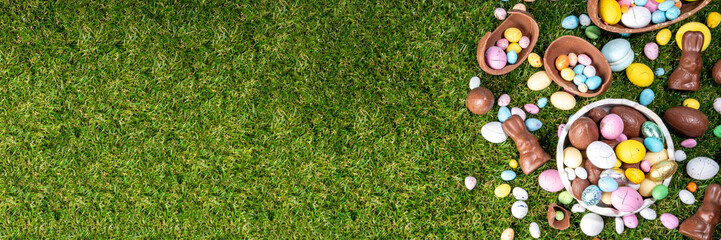 Fototapeta premium Easter egg hunting background with chocolate eggs. Various candy and chocolate Easter eggs, bunny and rabbits with basket for eggs on green grass park or garden background