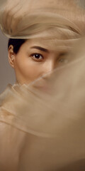 Asian woman beauty portrait behind translucent beige silk fabric revealing one eye. Luxury skincare...